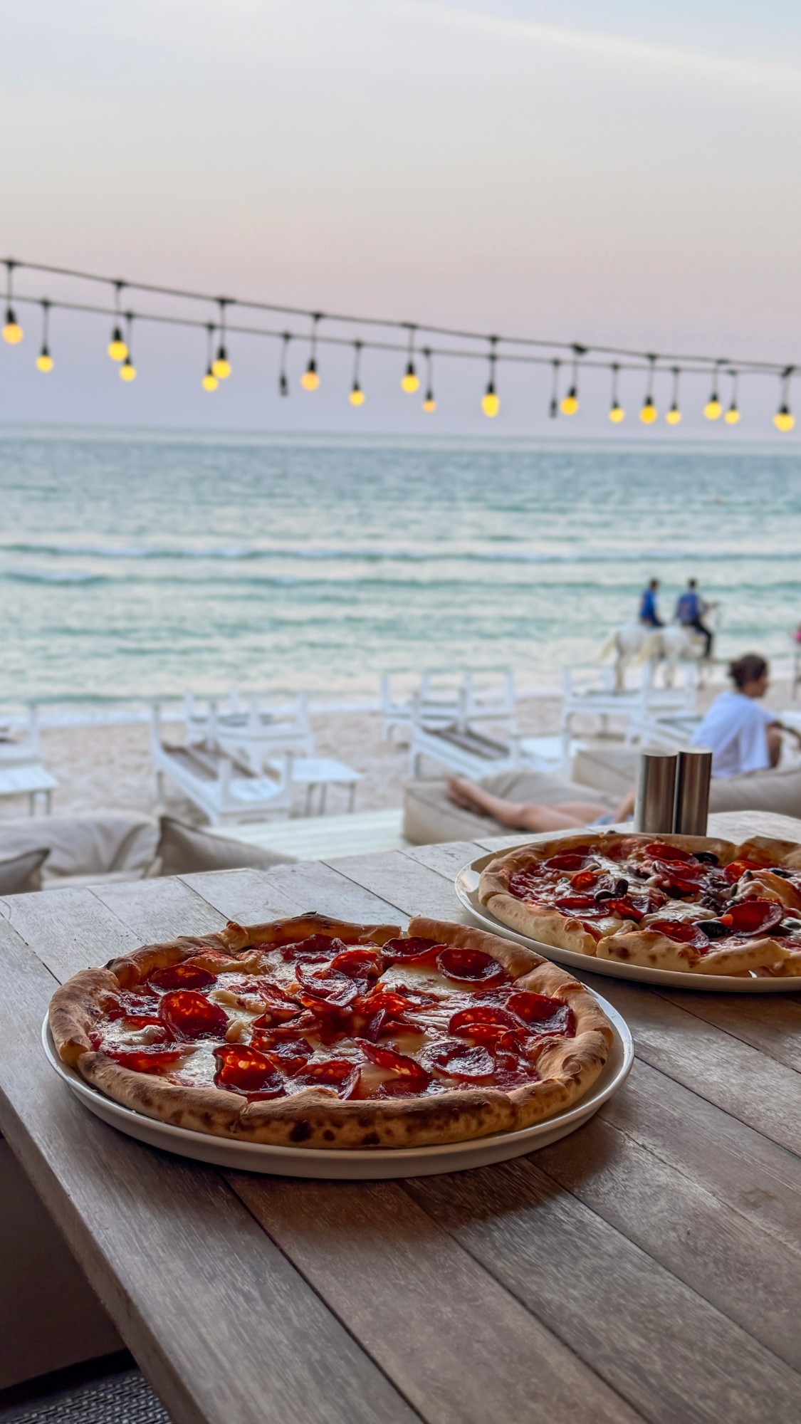 Two pizzas on a beachside table at sunset, sea and string lights behind